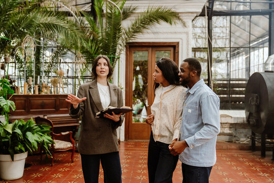 A woman and a man stand and hold hands while listening to a female organizer standing next to them and holding a clipboard while consulting