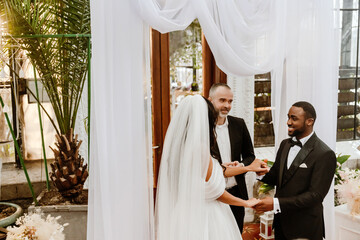 The priest stands and looks at the groom who stands next to him and smiles while holding the...