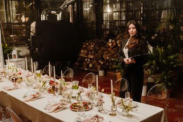 A woman organizer stands and holds a clipboard while looking at the table in front of her, in an establishment
