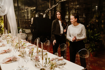 Female organizer holding clipboard while walking next to female waitress, they are looking at the table, in the establishment