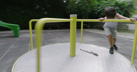 Boy spinning on a playground carousel, surrounded by green trees and a wooden fence in a serene park setting