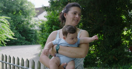 Fototapeta premium Mother holding baby outdoors, smiling as she engages with her child, with greenery and trees in the background, joyful and tender moment in a natural park setting