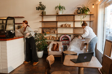 A female owner and a female employee stand and remove bags from equipment and furniture, in a cafe