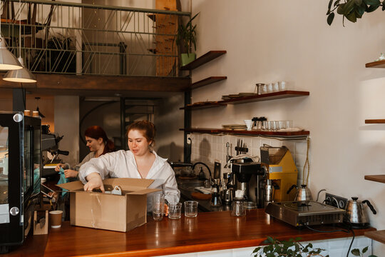 A female owner stands behind the bar and takes cups out of a box while a female employee stands behind her holding a towel, in a cafe - Powered by Adobe