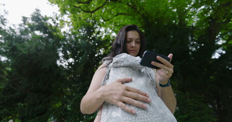 Woman multitasking in a park, holding her baby in a front carrier while focused on her smartphone,...