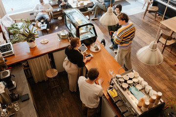 A female owner and a male employee stand behind the bar while a group of five customers sit and walk around next to them, in a cafe