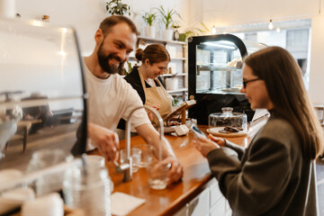A female owner stands behind the bar and looks at a computer tablet while a male employee stands next to her, pouring water into a glass while a female customer stands opposite him, in a cafe