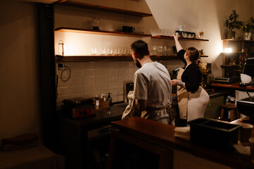 A female owner stands and places a cup on a shelf while a male employee stands next to her holding a towel, in a cafe