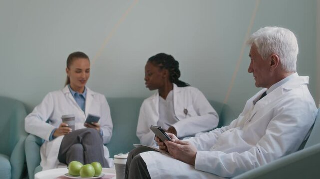 Side view of mature male physician watching video on digital tablet then greeting female colleagues casually talking on couch in background while resting in lounge area of clinic