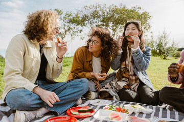 Group of four friends sitting on a blanket and talking while eating, at a picnic