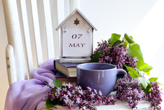 Calendar for May 7: numbers 07, word May in English on a decorative calendar, cup of tea, lilac branches on a chair