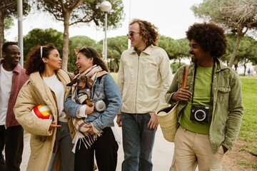 Group of five friends walking and laughing while talking and holding a ball and a dog, outdoors
