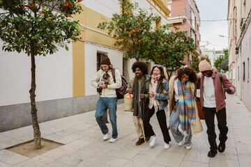 Fototapeta premium A group of four friends are walking and putting their arms around each other while a male friend walks next to them holding a dog, they are laughing, on the street