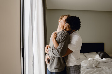 Fototapeta premium A man stands and hugs a man standing in front of him from behind, in a bedroom