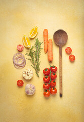 Cherry tomatoes, garlic, lemon slices, carrots, red onions, and rosemary sprig arranged alongside a wooden spoon on a yellow backdrop, culinary creativity and healthy eating concept