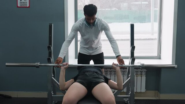 female client lying on bench press receiving spotting assistance from trainer under barbell lift with urban traffic visible through window with modern racks and sunlight