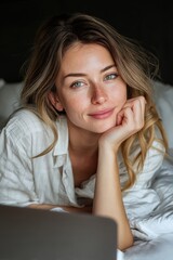 A woman with freckles and blond hair smiles while resting in bed with her laptop.