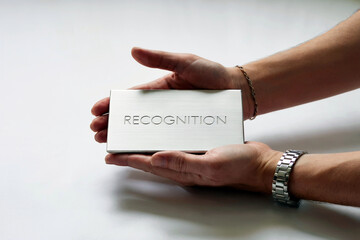 A pair of hands gently holds a sleek, metallic plaque engraved with the word "RECOGNITION," symbolizing achievement and appreciation against a clean, light background. By IA Generative