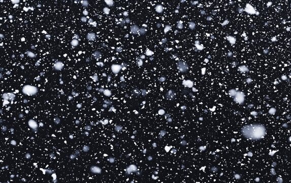 A close-up view of numerous snowflakes falling against a dark background, creating a winter scene.  The snowflakes vary in size and shape, with some appearing as delicate ice crystals