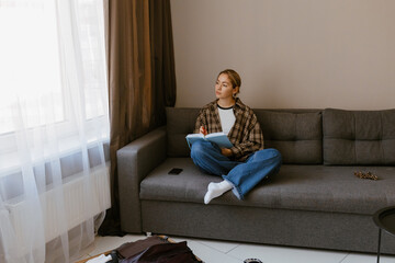 Obraz premium A woman is sitting on a sofa and looking away while holding a notebook, in the living room