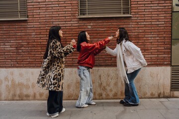 Naklejka premium A woman holds a bottle near the face of her female friend who is standing across from her while a female friend stands next to them, on the street