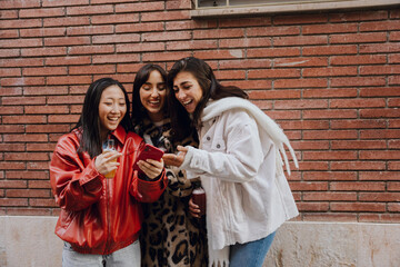 Group of three female friends standing and laughing while looking at a phone one of them is...
