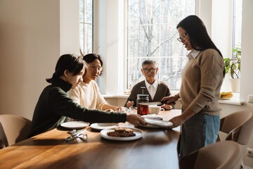 A grandmother stands and holds a knife while talking to her grandfather, son, and grandson who are sitting next to her at the table