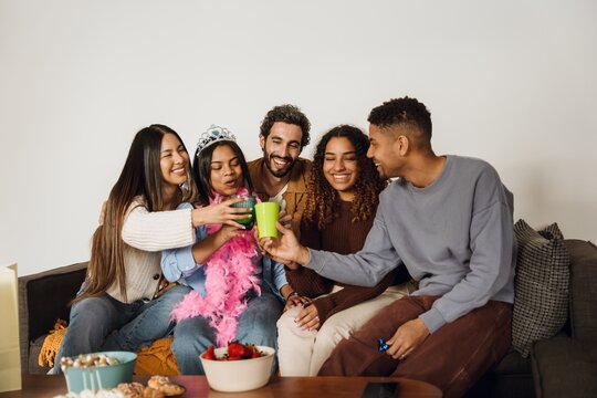 A group of five friends are sitting on a couch and smiling while clinking cups