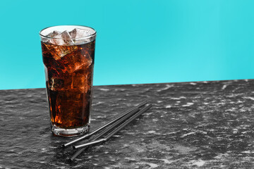 Refreshing cola with ice cubes in glass and straws on dark textured table against turquoise gradient background, closeup
