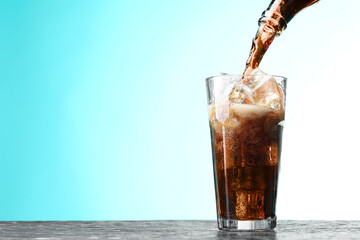 Pouring refreshing cola from bottle into glass at dark textured table against turquoise gradient background, closeup. Space for text