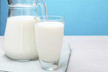 Fresh milk in glass and jug on white wooden table against light blue background, closeup. Space for text