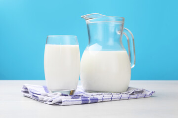 Fresh milk in glass and jug on white wooden table against light blue background