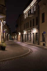 Obraz premium A vertical photo of a winding cobbled street in Vilnius Old Town at night. On both sides of the street are old buildings with illuminated windows and decorative lanterns on the walls. 