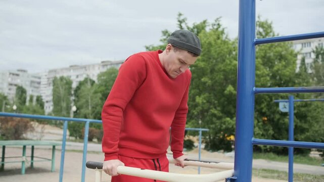 A man in a red outfit is engaging in strength training by performing dips on parallel bars in a city park. The urban environment features greenery and nearby buildings.