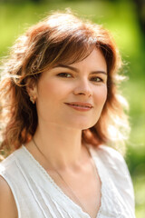 Fototapeta premium Close-up portrait of a beautiful woman with wavy brown hair, smiling softly while standing outdoors in natural sunlight. She is wearing a white blouse and minimal jewelry, with a calm expression. 