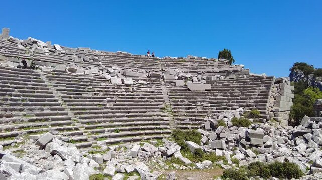 4K Tourists sitting on stone seats of the Termessos amphitheater, surrounded by towering mountains in the abandoned Pisidian city near Antalya, Turkey