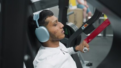 close up high school student sitting on exercise machine gripping handles while wearing wireless headset during gym workout session with other people casually seated in background
