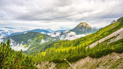 Majestic peaks rise above lush valleys in Low Tauern, Austria. Clouds drift lazily across the landscape, revealing patches of green and rocky terrain, creating a serene atmosphere.