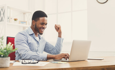 Fototapeta premium Happy excited businessman celebrate his success. Winner, black man in office reading on laptop, copy space