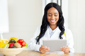 Happy African American nutritionist working with tablet computer, giving online consultation at...