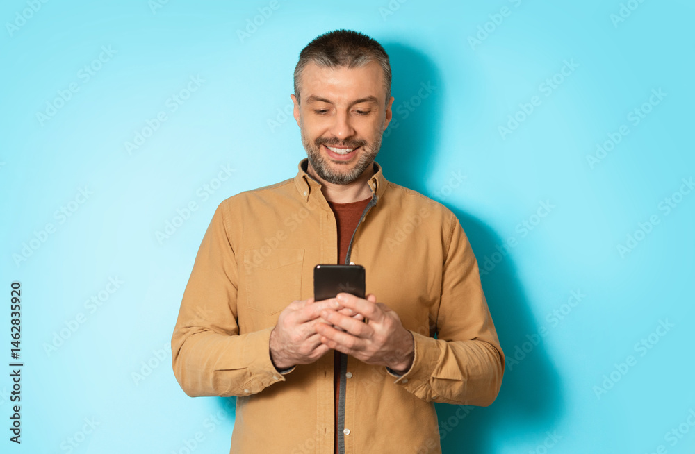 Poster Cheerful Man Using Mobile Application On Cellphone Or Texting Via Smartphone Communicating Online Standing In Studio Over Blue Background. Communication And Technology