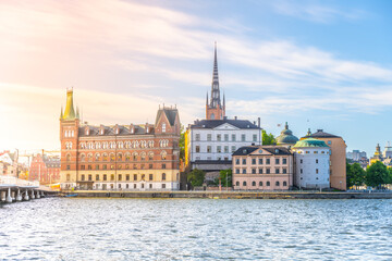 Naklejka premium Riddarholmen Island showcases beautiful architecture along the water's edge in Stockholm. The sun casts gentle light, highlighting historic buildings and a serene atmosphere.