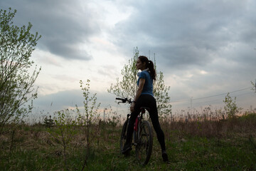 Young sporty woman riding a bicycle on the spring field with green grass on sunset sky landscape...