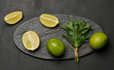 Whole and cut limes with fresh mint leaves arranged on a dark textured surface.