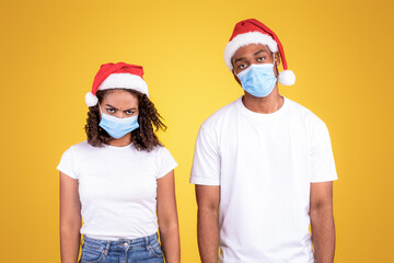 Fototapeta premium Coronavirus Christmas. Frustrated African American Couple Wearing Santa Hats And Protective Medical Masks Looking At Camera Standing On Yellow Studio Background. Xmas Party Cancelled Due To Covid-19