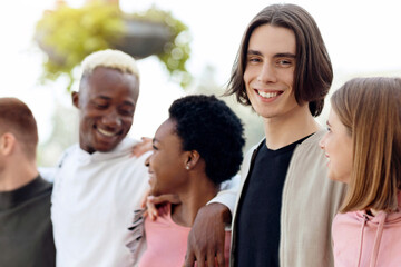 Cheerful handsome guy spending time with his international friends men and women, walking by street and embracing, having conversation, smiling and laughing, panorama. Millennials lifestyle
