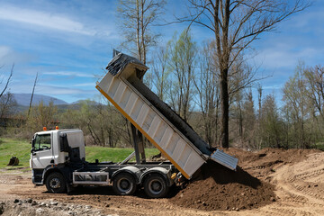 White dump truck unloading soil at a construction site. Heavy machinery performing earth works, backfilling for a landscape architecture project. Industrial earthmoving operation. © Christos