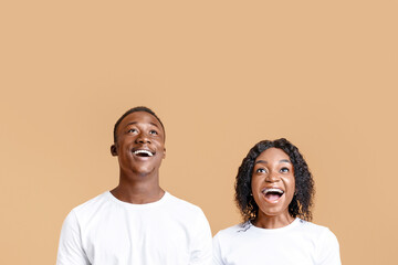 Fototapeta premium Excited black couple looking up at copy space over yellow studio background. Amazed african american man and woman watching advertisement or announcement about seasonal sales and offers