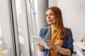 Female business expert with tablet in hands looking through the window while standing at corporate office.