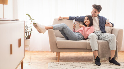 Asian Millennial Couple Using Smartphones Relaxing Sitting On Couch At Home On Weekend. Gadget...